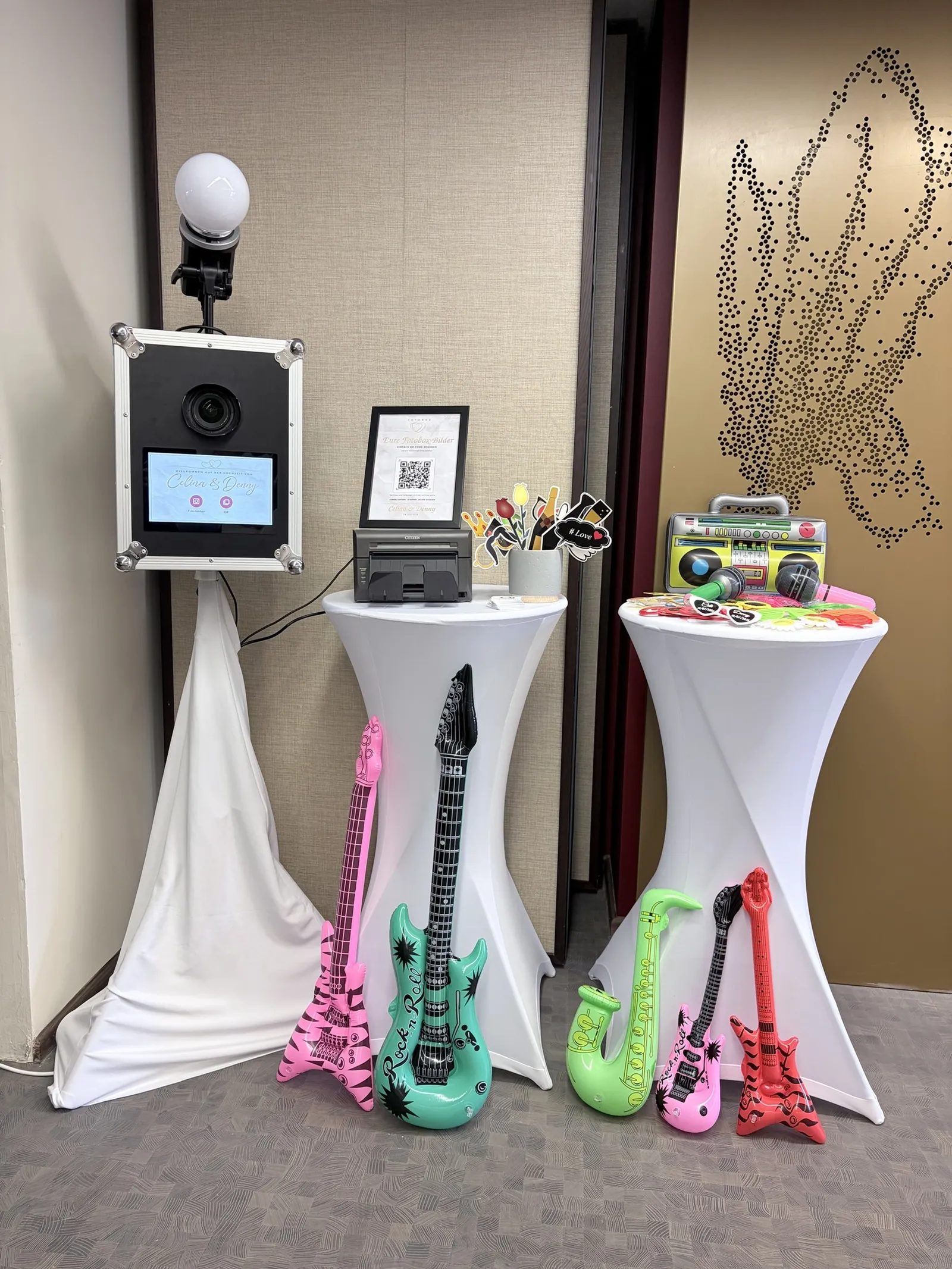 Fotobox mit Studioblitz und Touchscreen, daneben Drucker und Stehtische mit Hussen, aufblasbare Gitarren und weitere Accessoires