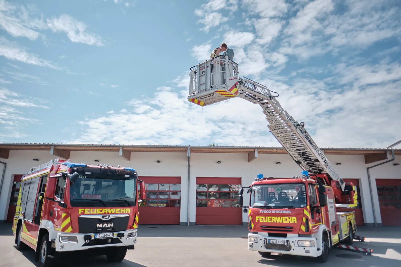 Brautpaar winzig vor der ausgefahrenen Drehleiter zwischen zwei Feuerwehrfahrzeugen – Hochzeit Ronja & Markus, Feuerwehrhaus Buchloe