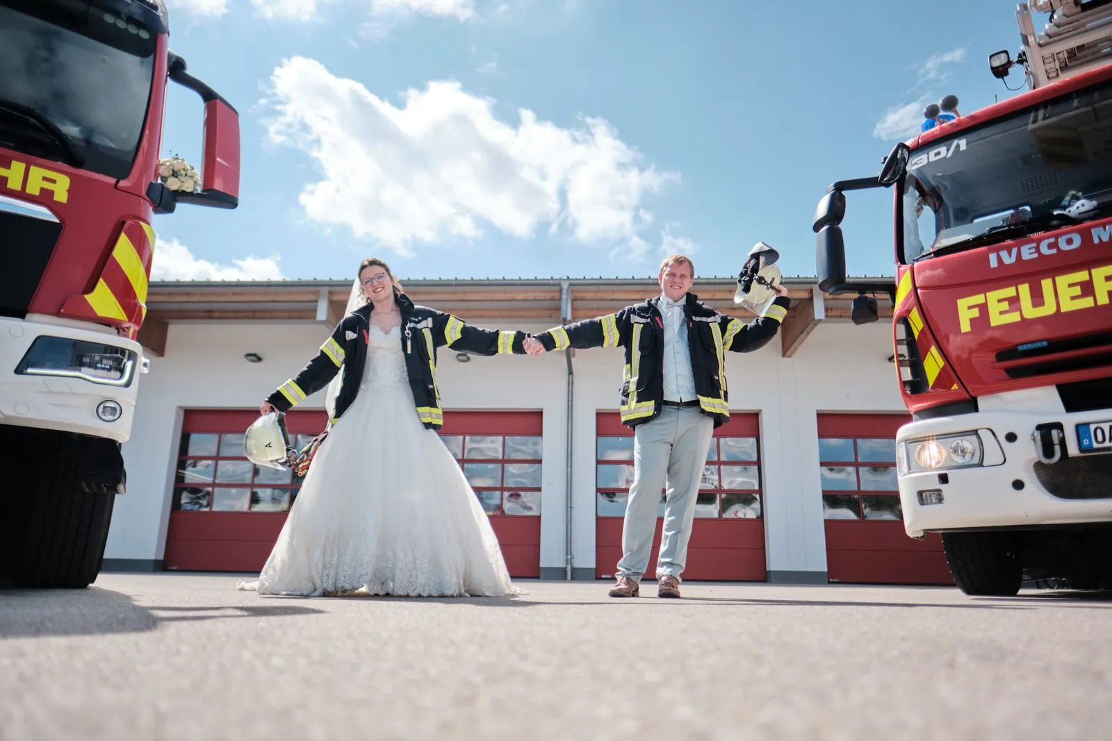 Ronja und Markus halten ein Blumenband zwischen sich, Feuerwehrfahrzeuge im Hintergrund – Hochzeit Ronja & Markus, Feuerwehrhaus Buchloe