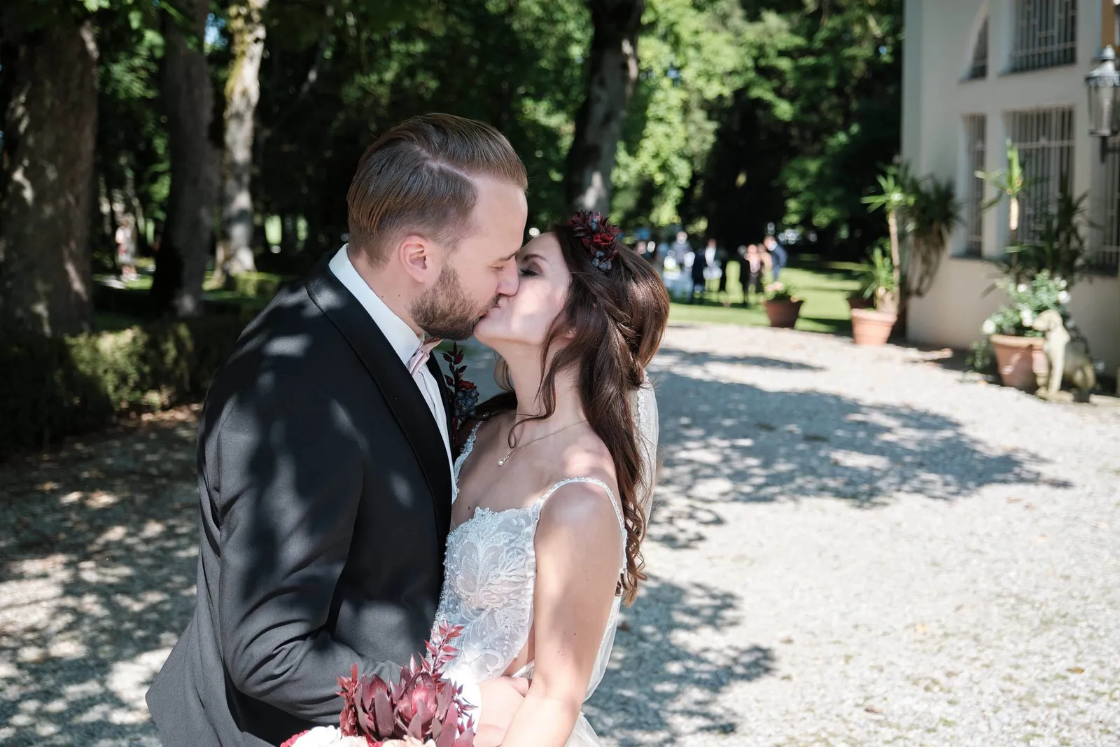 Brautpaar küsst sich auf einem Kiesweg im Schatten der Bäume – Hochzeit Charly & Moritz