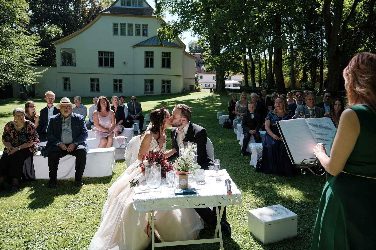 Brautpaar küsst sich am Trautisch, Gäste sitzen im Garten – Hochzeit Charly & Moritz, Herrenhaus Bannacker