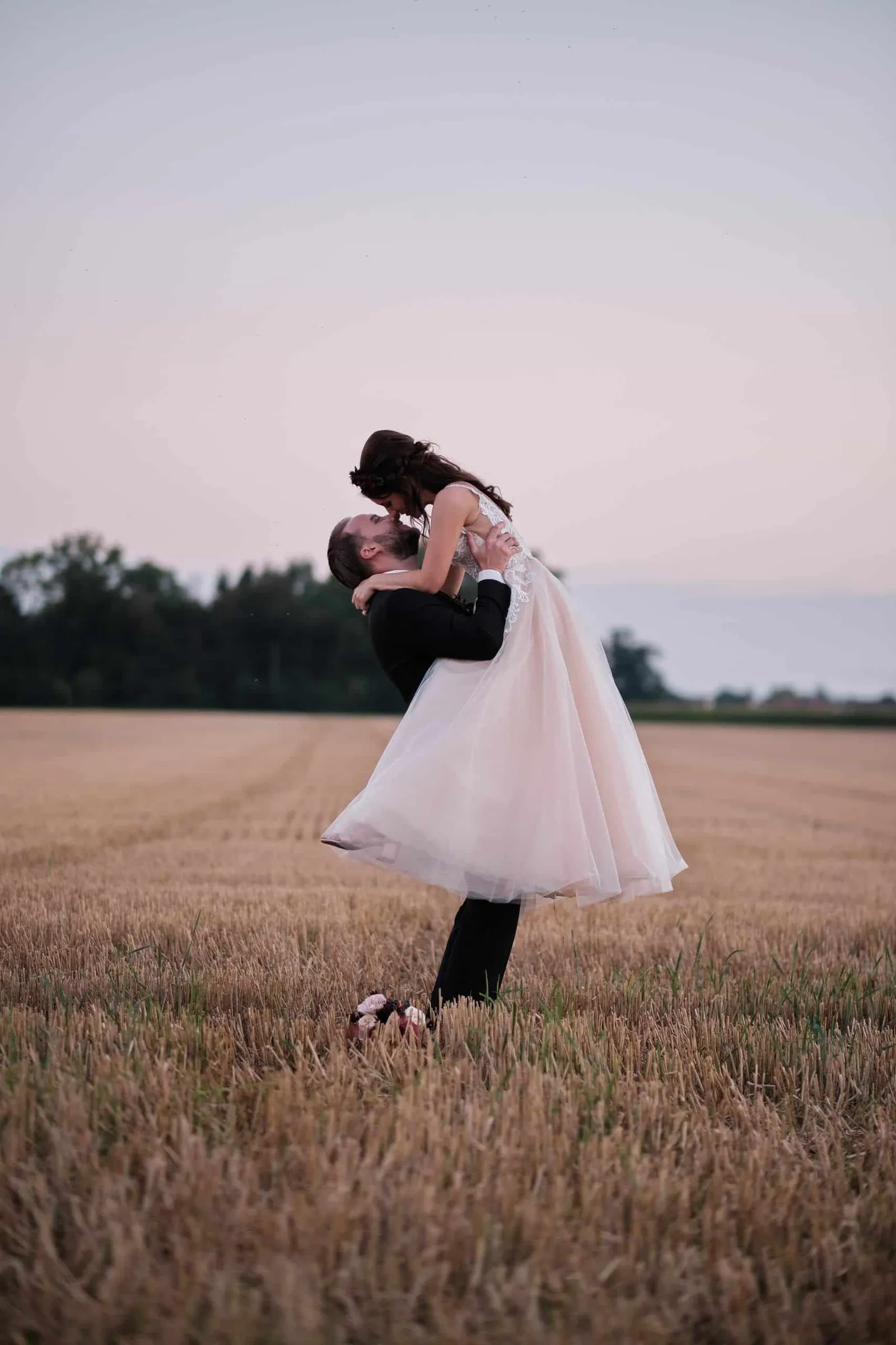 Bräutigam hebt die Braut in der Abenddämmerung im Stoppelfeld hoch – Hochzeit Charly & Moritz