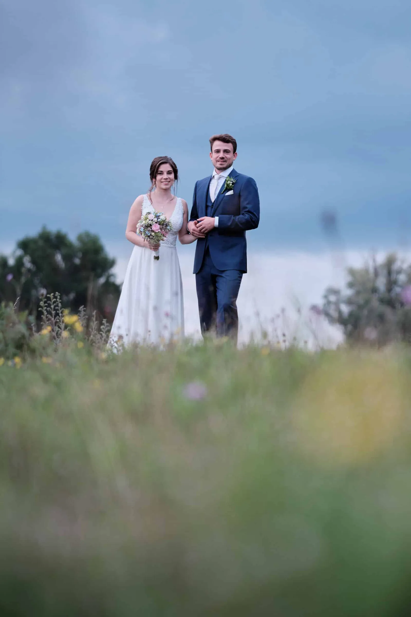 Brautpaar steht Arm in Arm auf einer Wiese, Gräser im Vordergrund – Hochzeit Hanna & Tobi