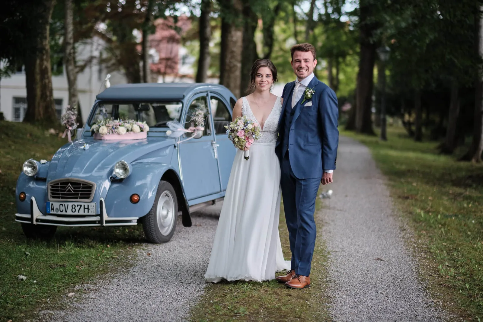 Brautpaar vor einem hellblauen Citroën 2CV auf einem Kiesweg – Hochzeit Hanna & Tobi
