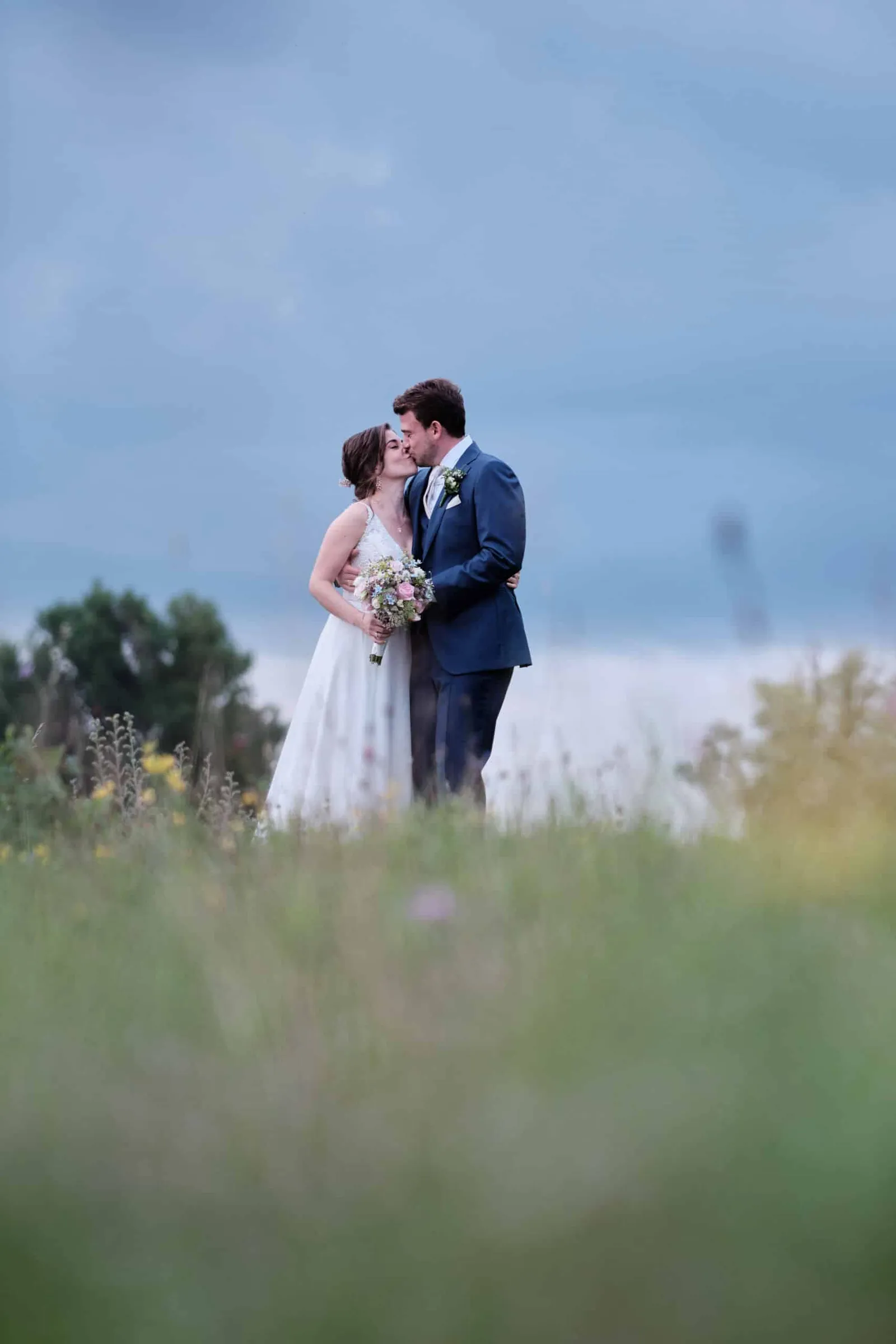 Brautpaar küsst sich auf einer Wiese unter bewölktem Himmel – Hochzeit Hanna & Tobi