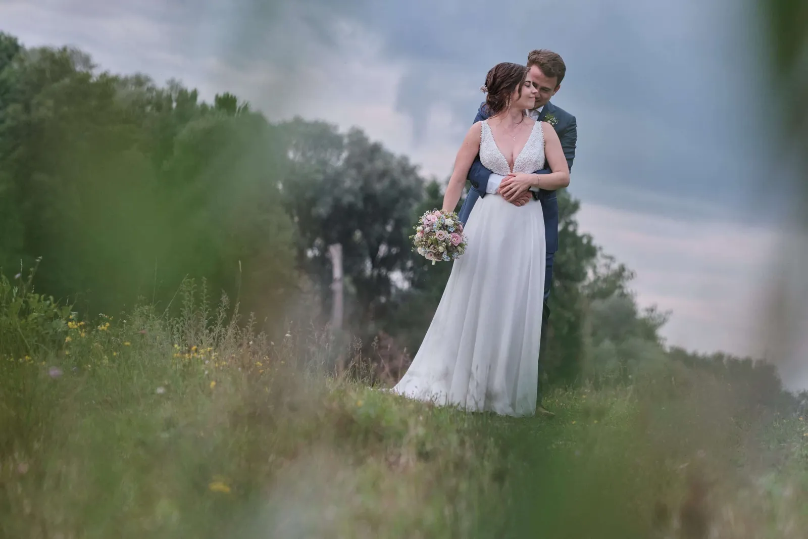 Bräutigam umarmt die Braut von hinten, unscharfes Grün im Vordergrund – Hochzeit Hanna & Tobi
