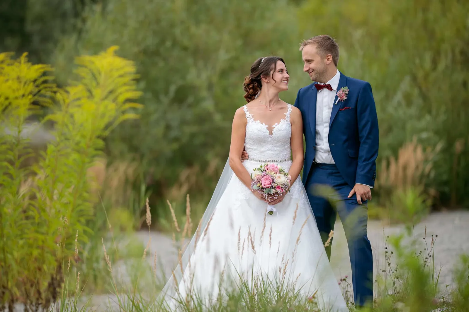 Brautpaar mit Brautstrauß zwischen hohen Gräsern und gelben Blumen – Hochzeit Julia & Phil