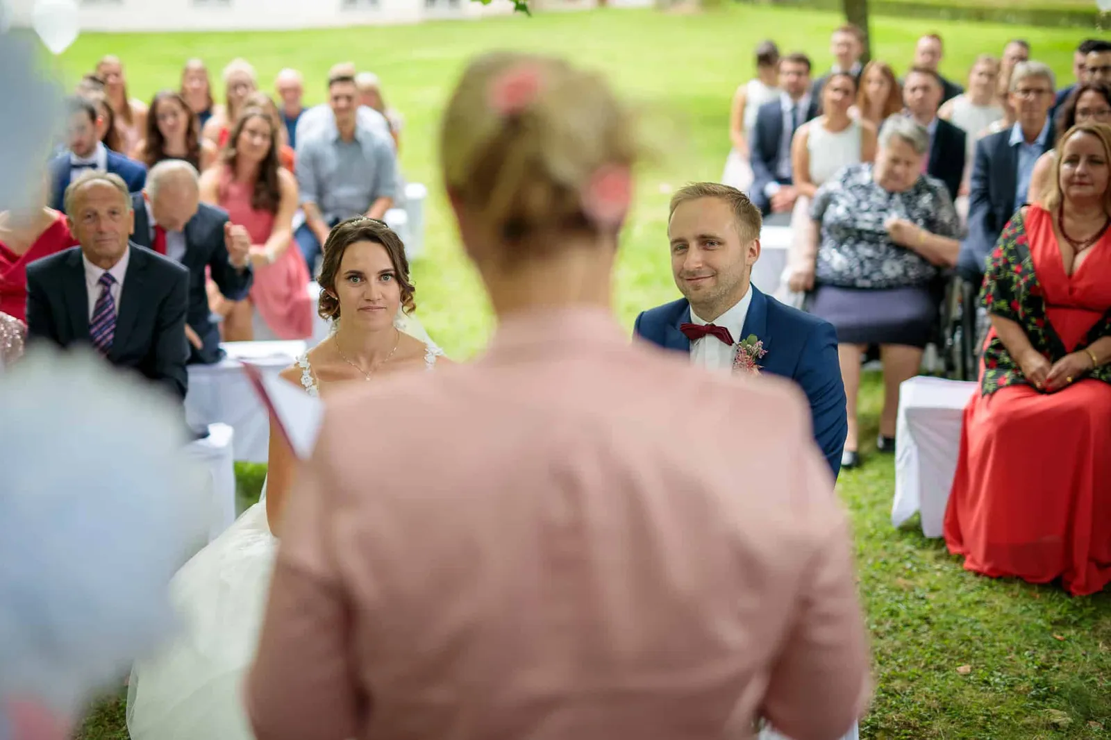 Traurednerin von hinten, Brautpaar und Gäste sitzen im Garten – Hochzeit Julia & Phil, Herrenhaus Bannacker