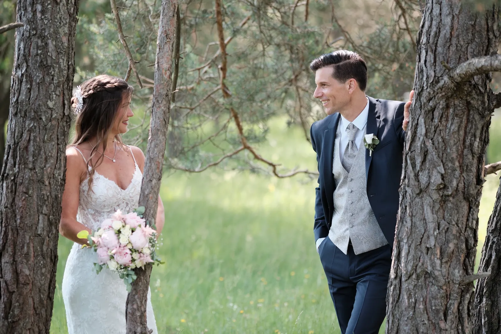 Brautpaar lugt lachend hinter einem Kiefernstamm hervor – Hochzeit Markus & Tanja, Königsbrunner Heide