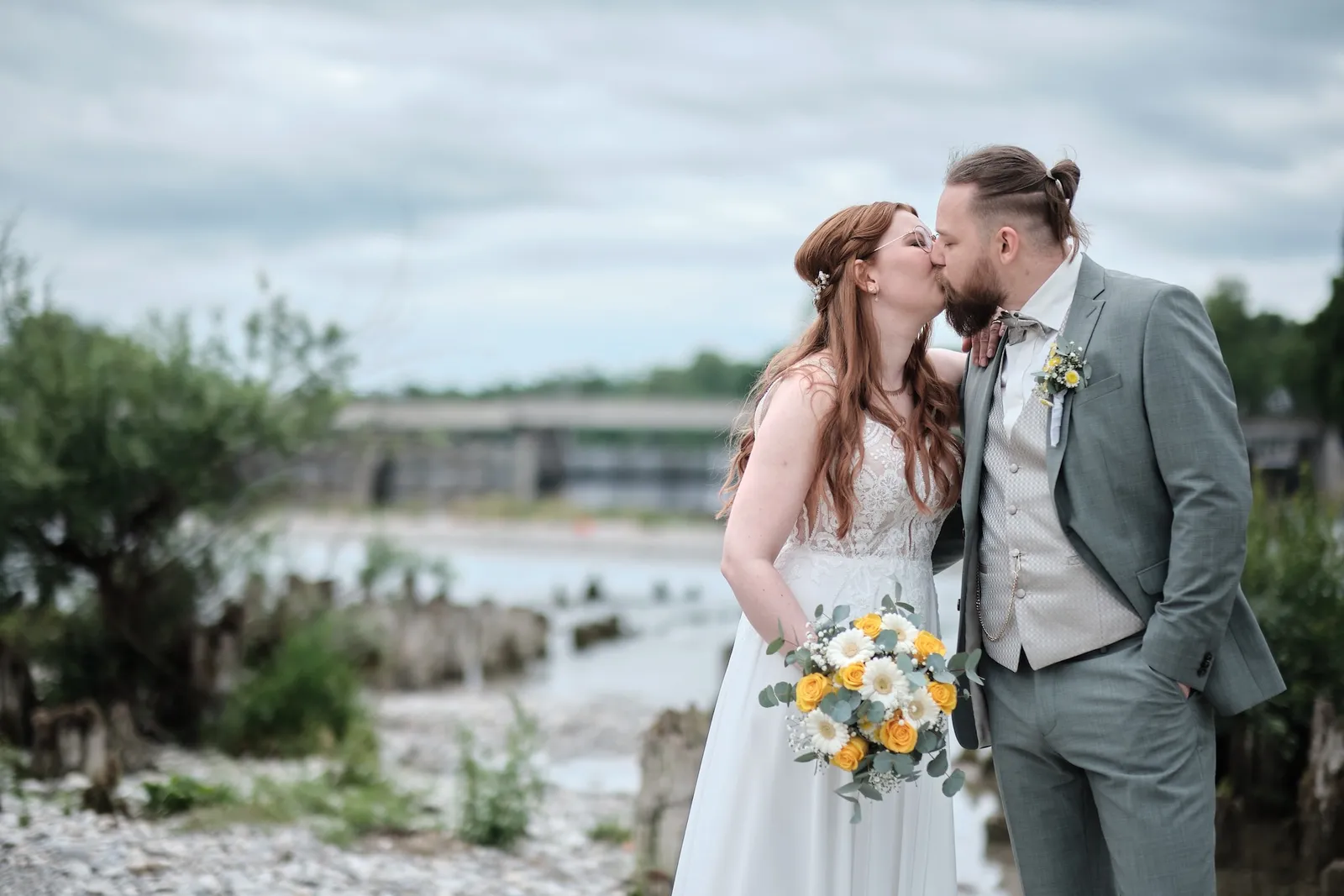 Brautpaar küsst sich am Lechufer, im Hintergrund das Wehr am Hochablass – Hochzeit Svenja & Markus