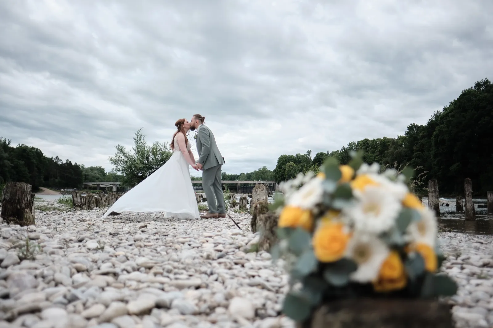 Brautstrauß im Vordergrund, dahinter küsst sich das Brautpaar auf der Kiesbank – Hochzeit Svenja & Markus