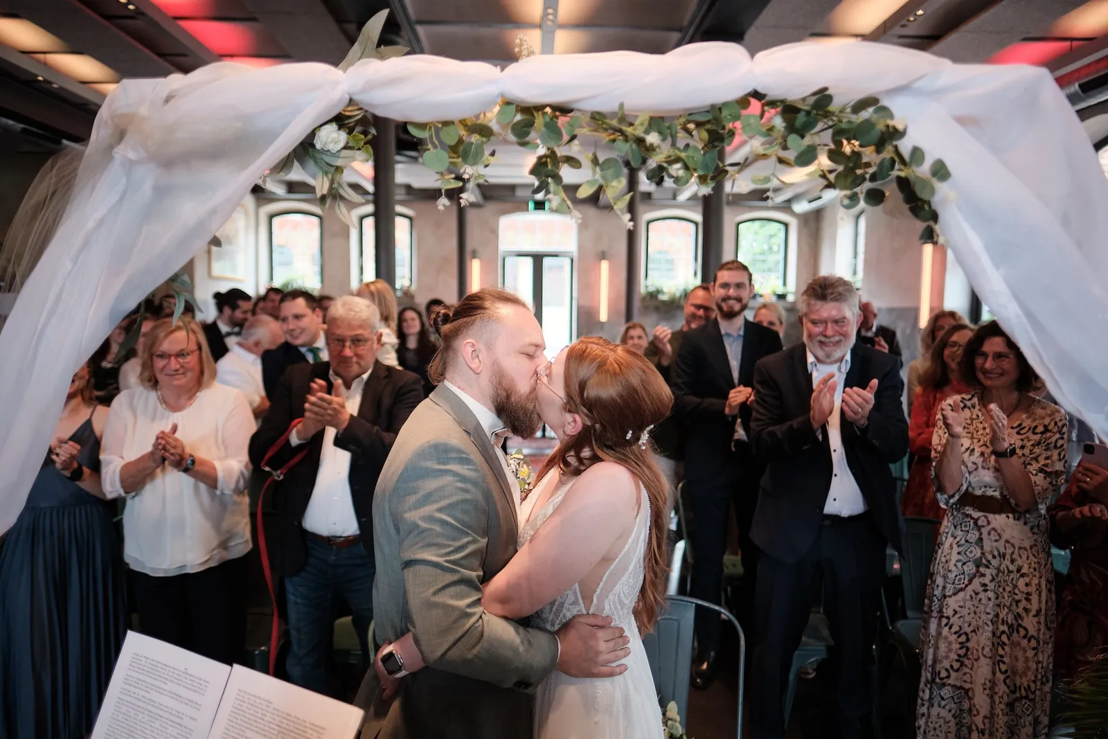 Erster Kuss als Ehepaar unter dem Traubogen, die Gäste applaudieren – Hochzeit Svenja & Markus, N8Stallung Augsburg
