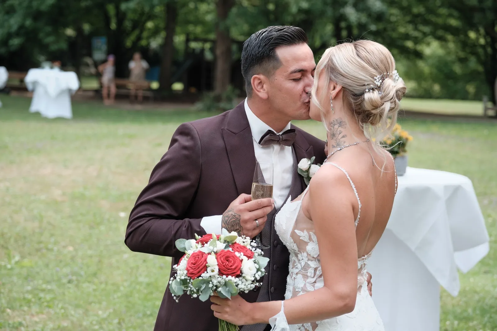Brautpaar küsst sich im Park, Celina hält den Brautstrauß mit roten Rosen – Hochzeit Celina & Denny, Parkhotel Schmid Adelsried
