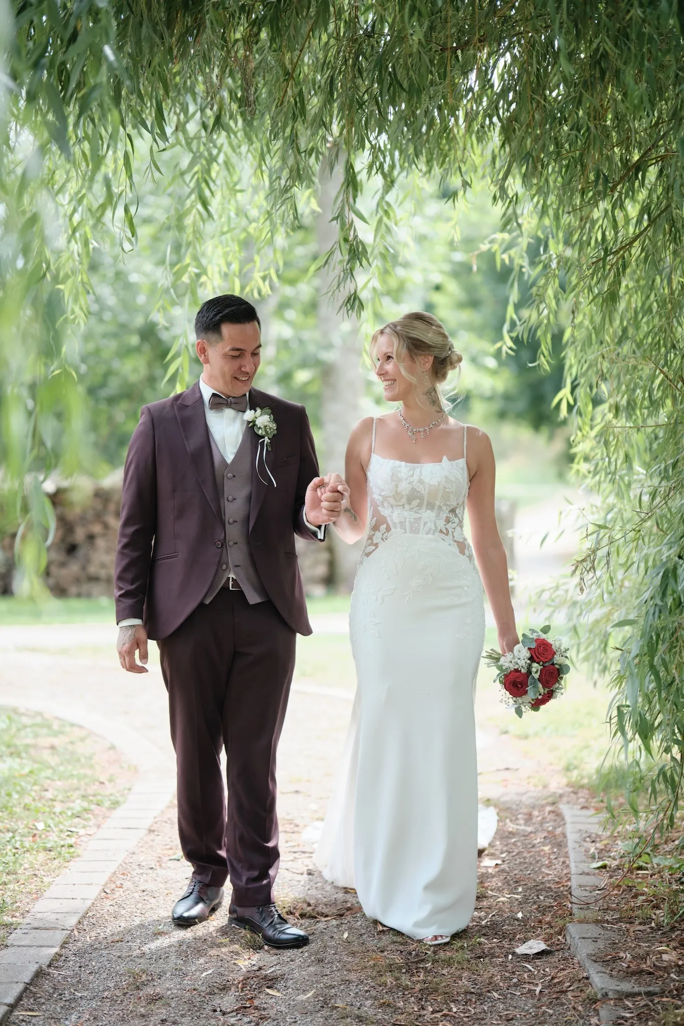Brautpaar geht Hand in Hand unter einer Trauerweide im Park, Celina mit Brautstrauß aus roten Rosen – Hochzeit Celina & Denny, Parkhotel Schmid Adelsried