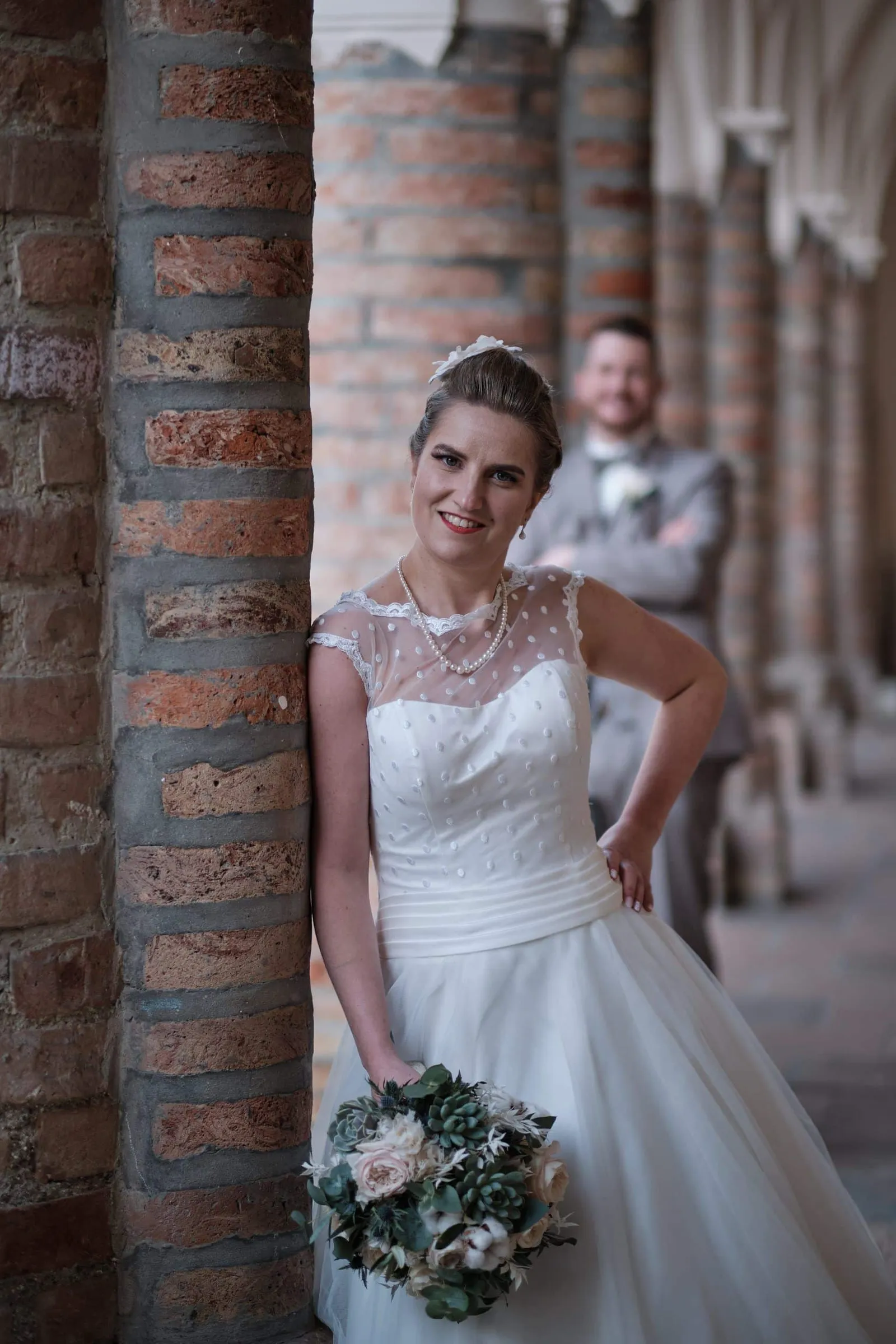 Lächelnde Braut an einer Backsteinsäule, Bräutigam im Hintergrund – Hochzeit Andi & Betti