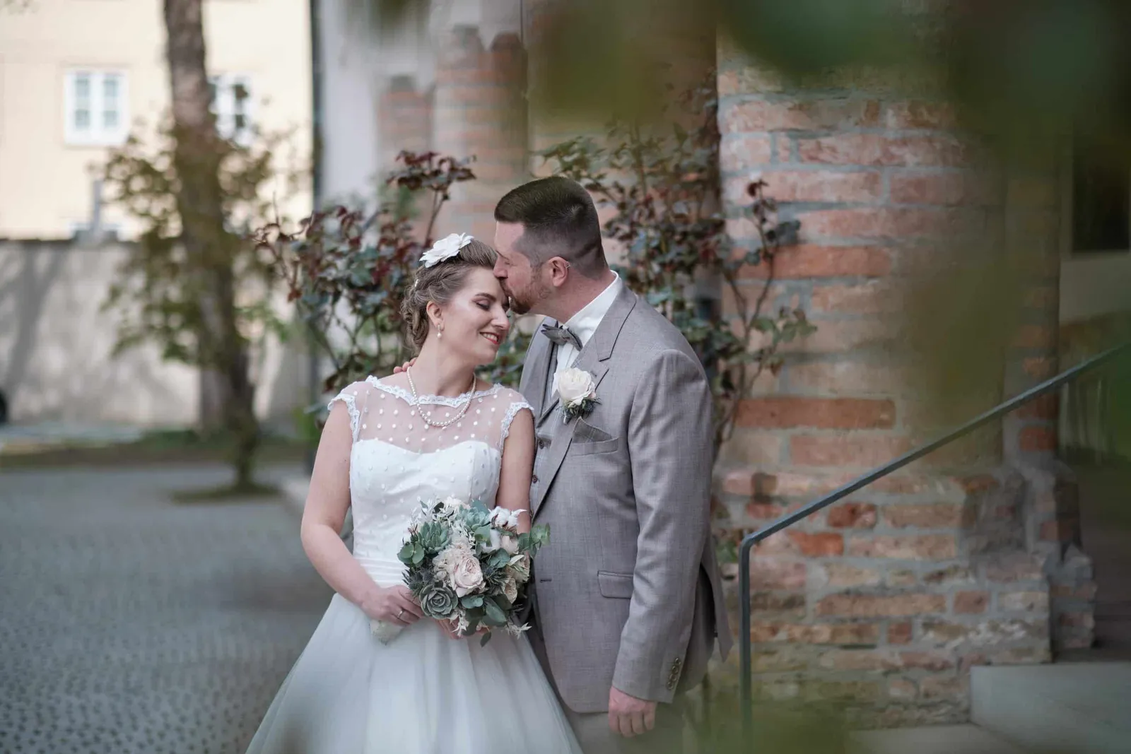 Brautpaar Stirn an Stirn mit Brautstrauß vor einer Backsteinwand – Hochzeit Andi & Betti