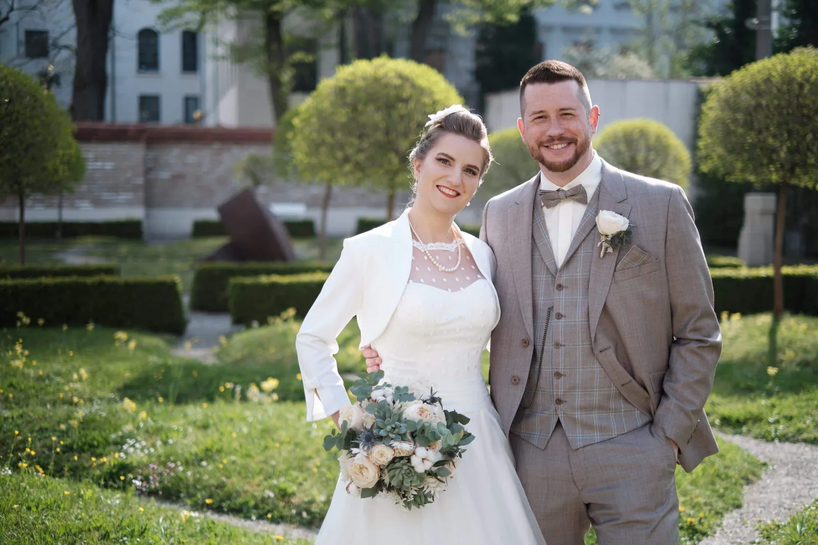 Lachendes Brautpaar mit Strauß in einem Garten mit Buchskugeln – Hochzeit Andi & Betti