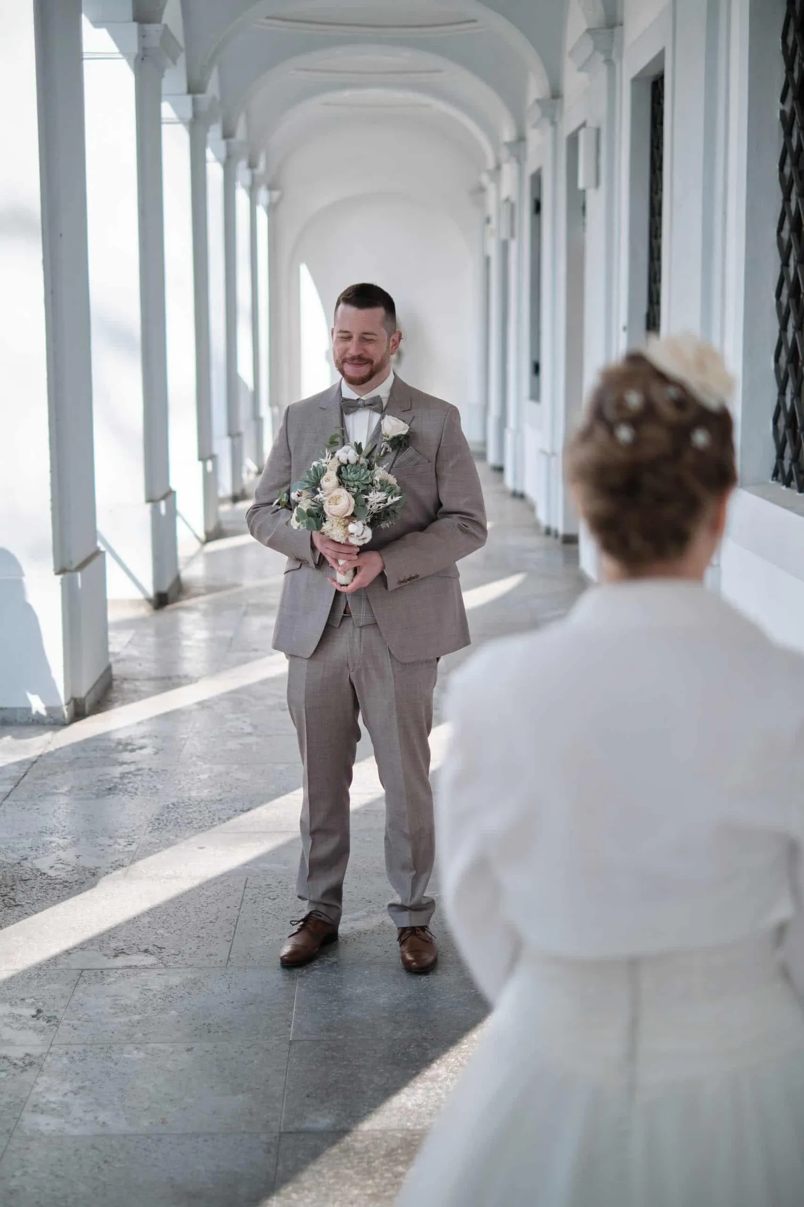 First Look: Bräutigam mit Strauß wartet im Arkadengang auf die Braut – Hochzeit Andi & Betti