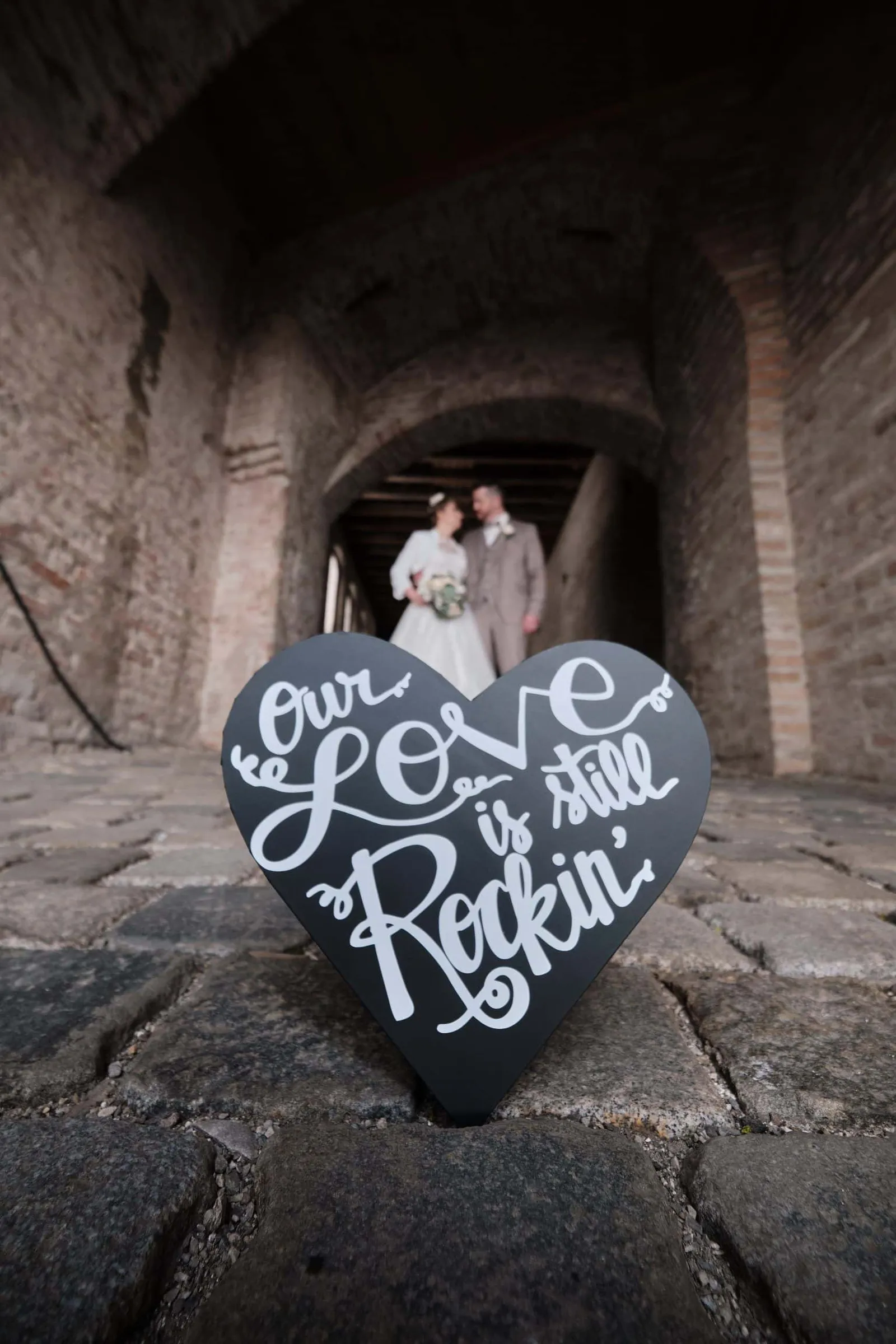 Herzschild „Our Love is still Rockin“, Brautpaar im Torbogen dahinter – Hochzeit Andi & Betti, Schaezlerpalais