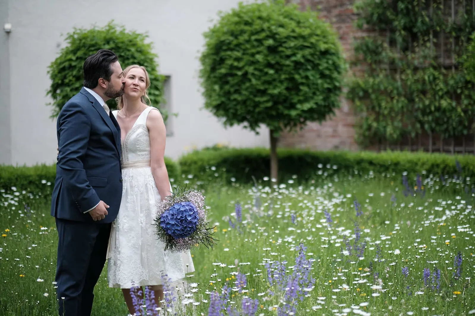Bräutigam küsst die Braut auf die Wange auf einer Blumenwiese – Hochzeit Katharina & Christian