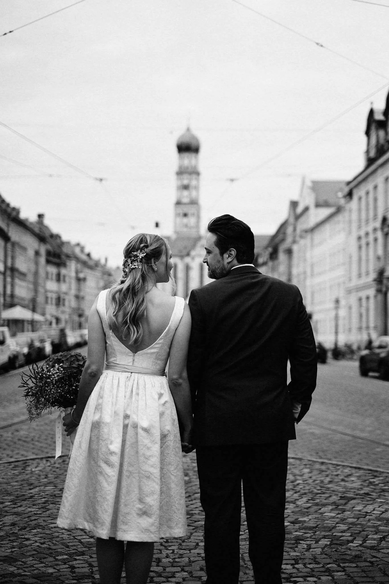 Brautpaar Hand in Hand von hinten auf einer Straße mit Kirchturm – Hochzeit Katharina & Christian
