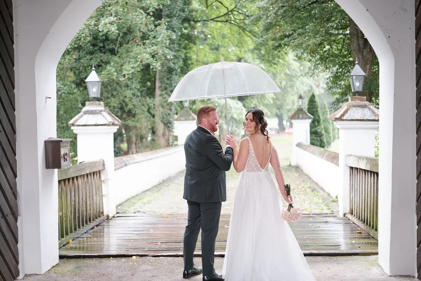 Brautpaar unter einem durchsichtigen Regenschirm auf der Zufahrt zum Schloss – Hochzeit Barbara & Christian, Schloss Neuburg a.d. Kammel