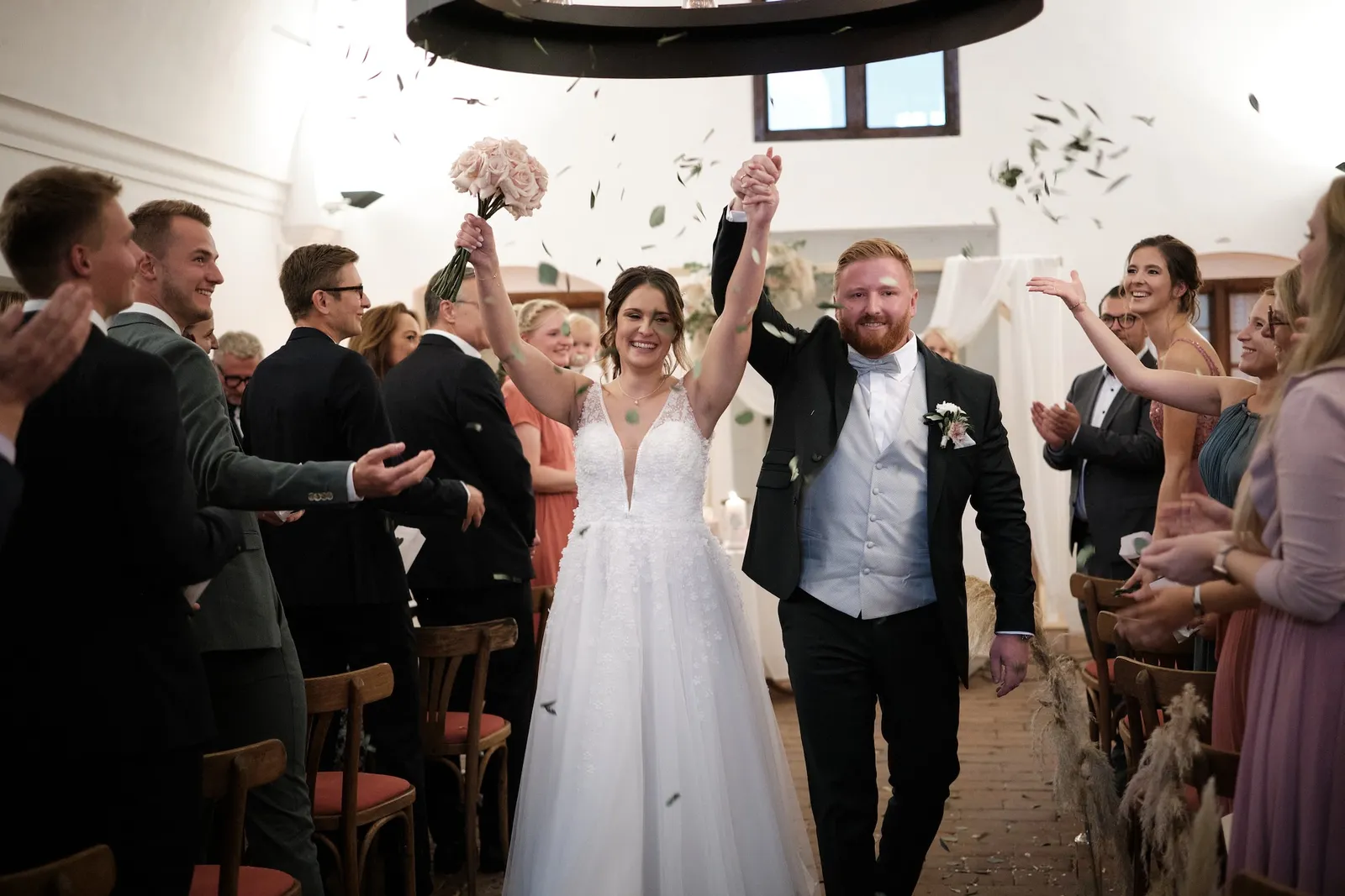 Brautpaar zieht mit erhobenen Händen durch Konfetti aus dem Gewölbe aus – Hochzeit Barbara & Christian, Schloss Neuburg a.d. Kammel