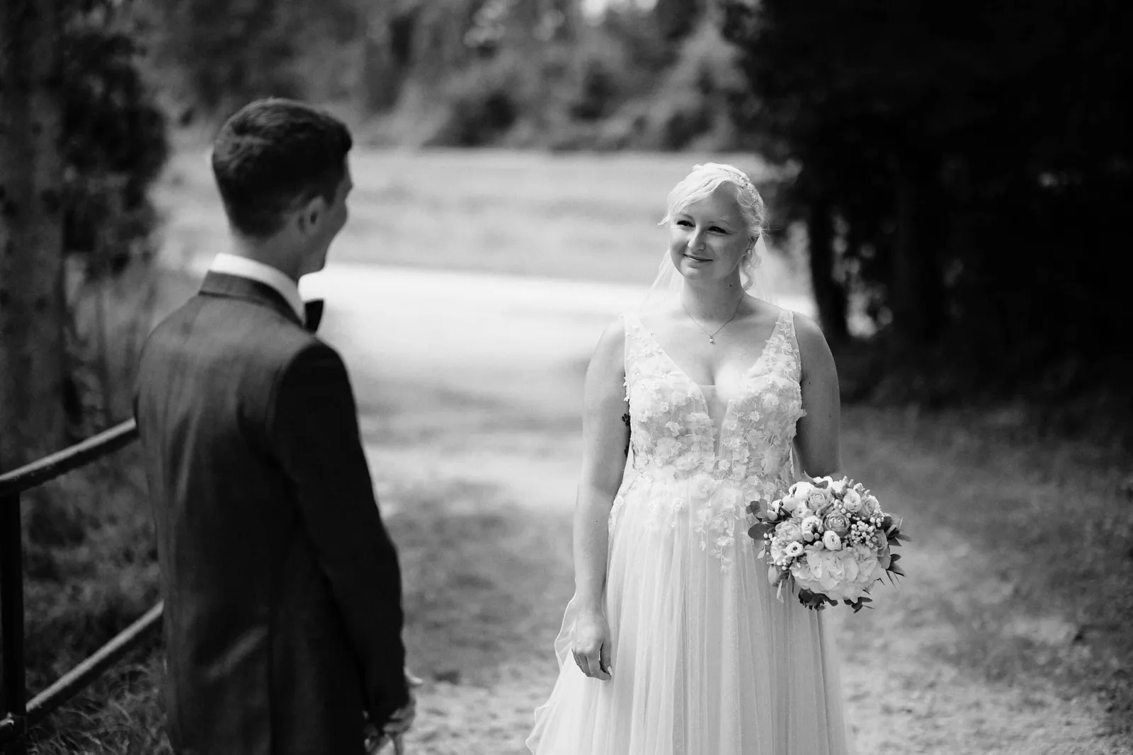 Braut mit Strauß lächelt dem Bräutigam auf einem Waldweg zu, schwarzweiß – Hochzeit Nina & Kevin