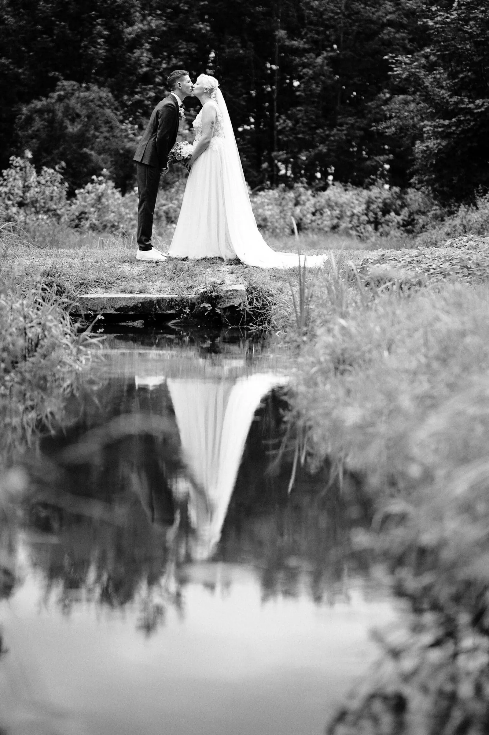 Brautpaar küsst sich am Teich und spiegelt sich im Wasser, schwarzweiß – Hochzeit Nina & Kevin
