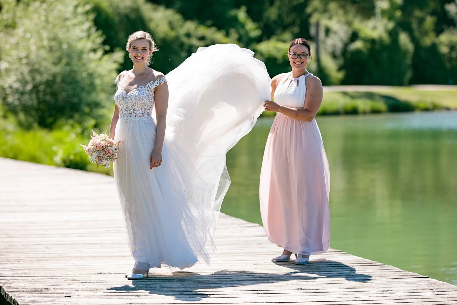 Braut mit Brautjungfer im rosa Kleid, die die Schleppe hochhält, auf dem Steg – Hochzeit Andreas & Nicole, Weitmannsee