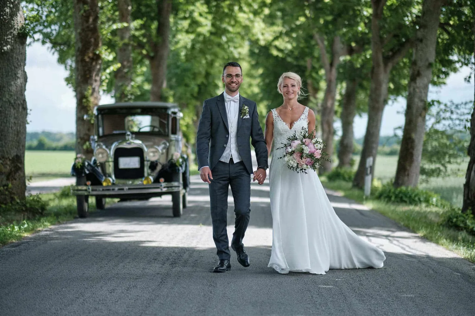 Brautpaar geht Hand in Hand vor einem Oldtimer durch eine Baumallee – Hochzeit Jessie & Kosta