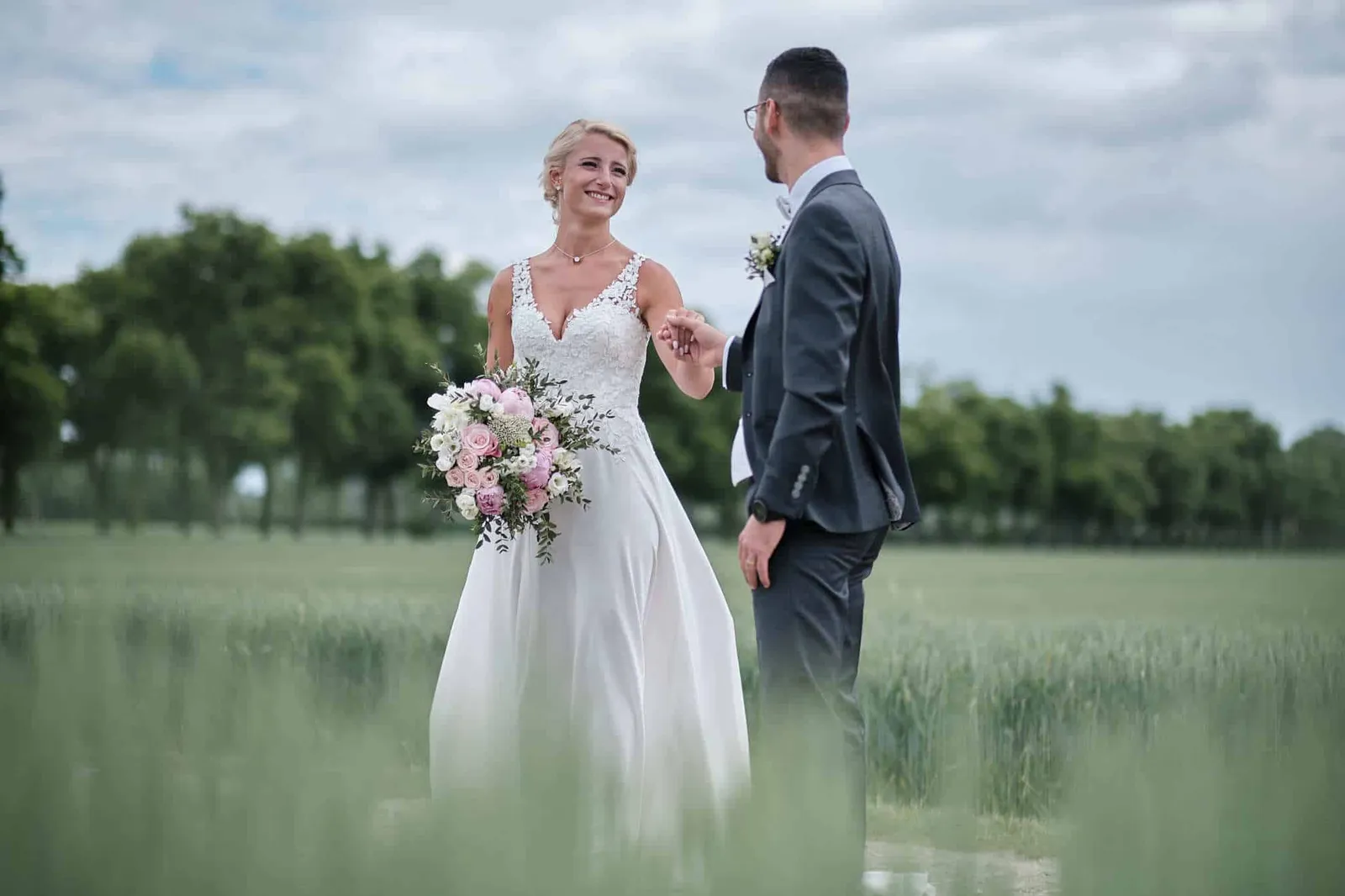 Braut mit Strauß lacht, Bräutigam hält ihre Hand im Getreidefeld – Hochzeit Jessie & Kosta