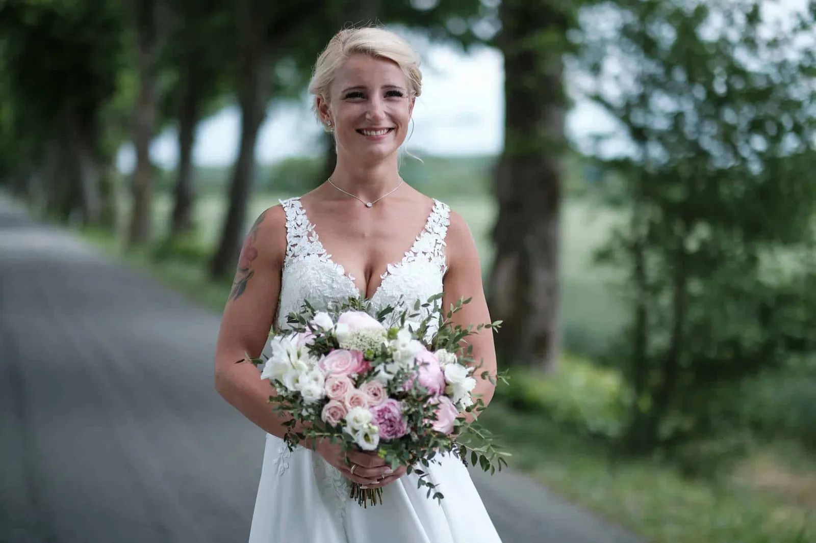 Lächelnde Braut mit Brautstrauß in einer Baumallee – Hochzeit Jessie & Kosta