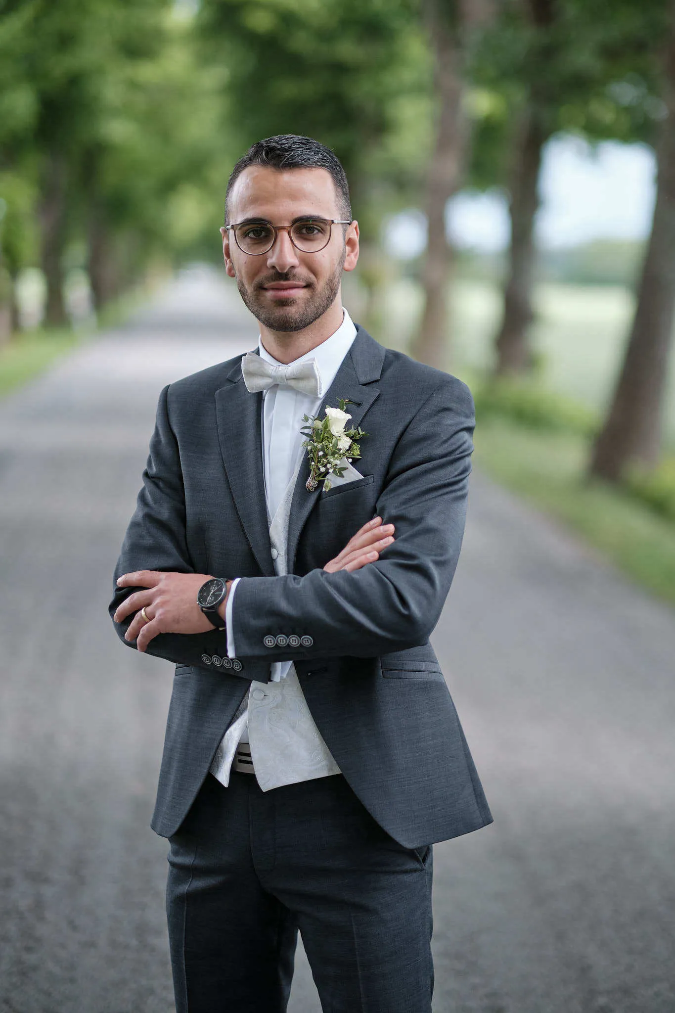 Bräutigam mit Brille und verschränkten Armen in einer Baumallee – Hochzeit Jessie & Kosta