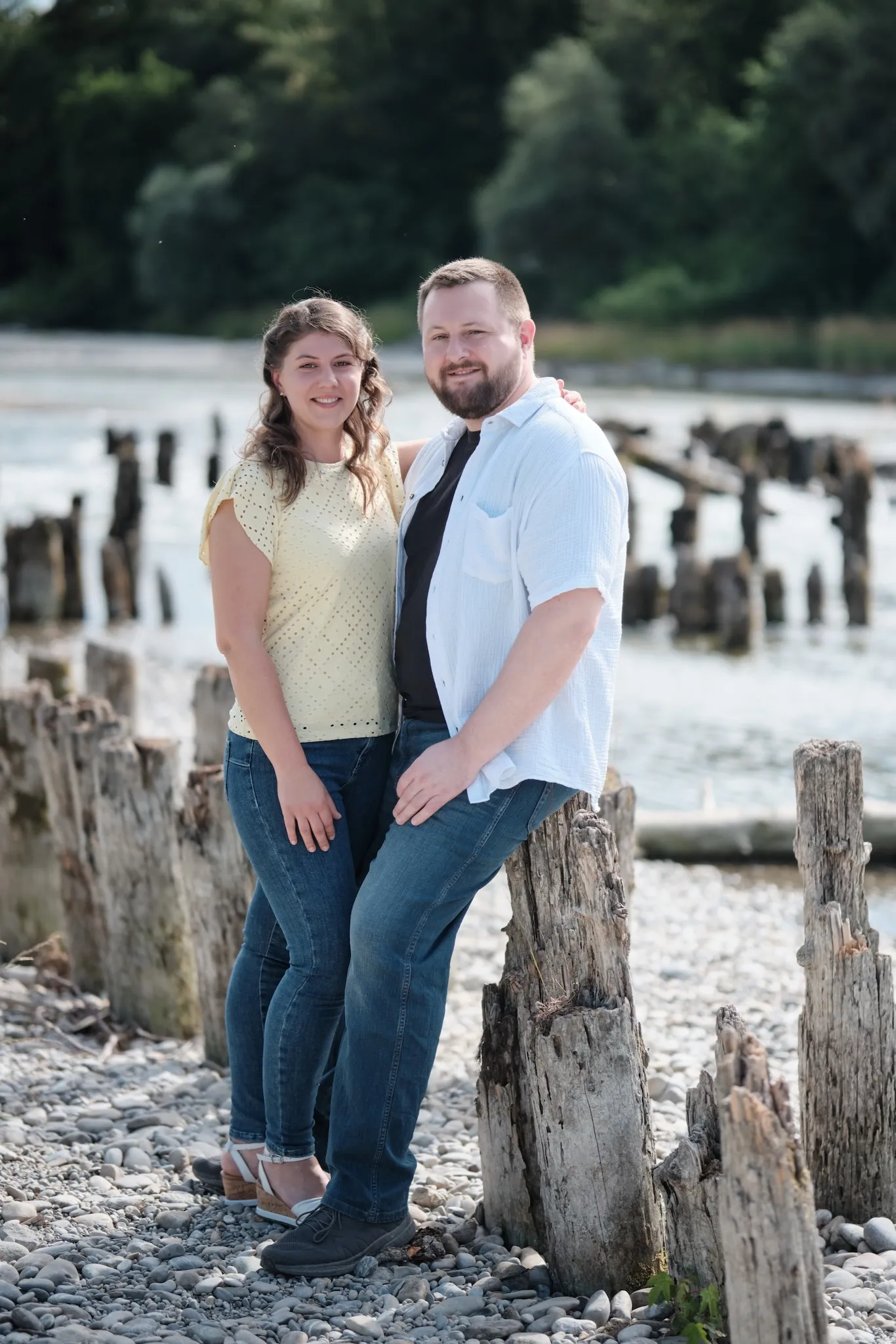 Umarmung an einem alten Holzpfahl an der Kiesbank, grüner Uferwald im Hintergrund – Kennenlernshooting Matthias & Rebecca, Hochablass Augsburg