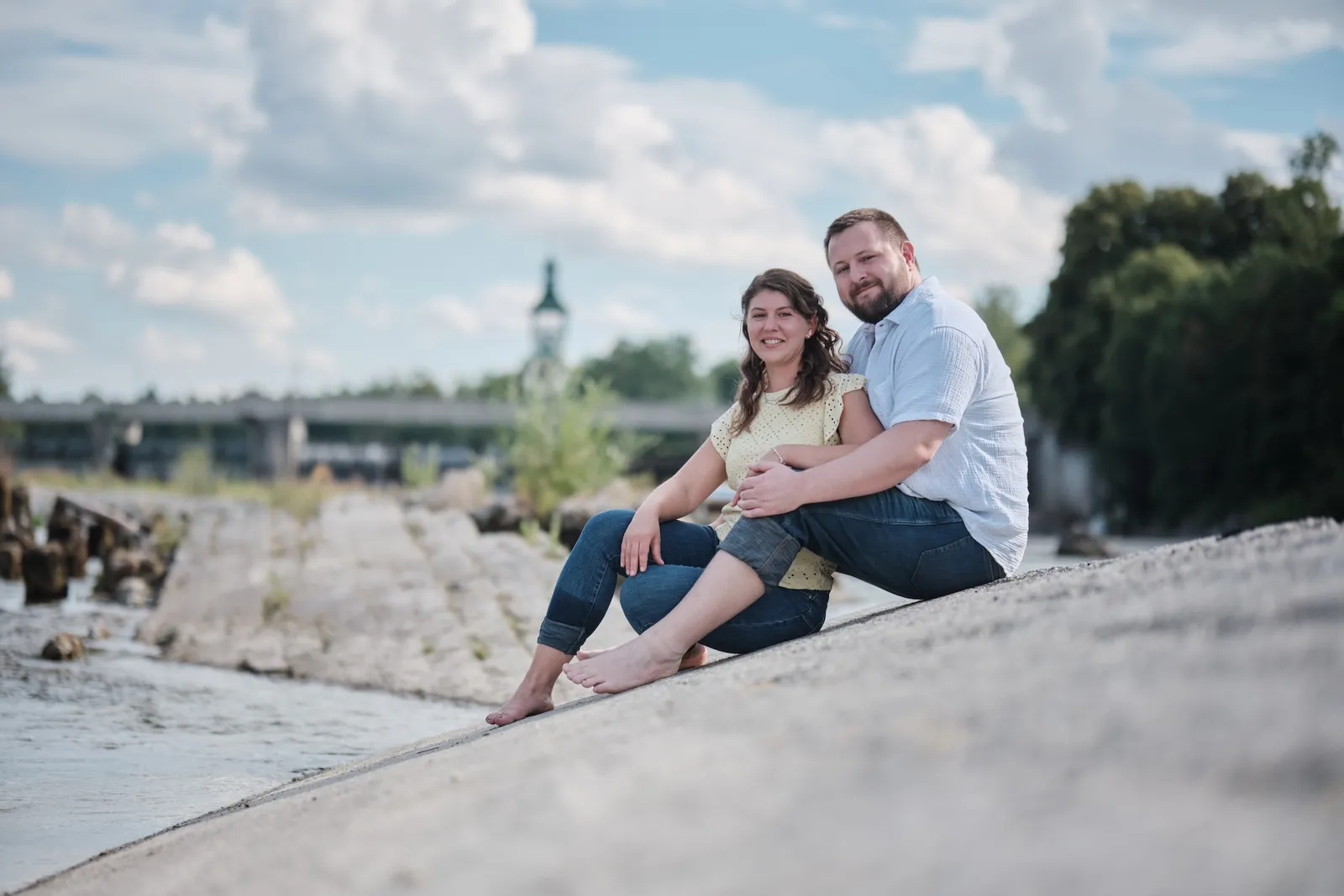 Vertraut nebeneinander sitzend auf der Betonschräge am Lech, Brücke im Hintergrund – Kennenlernshooting Matthias & Rebecca, Hochablass Augsburg