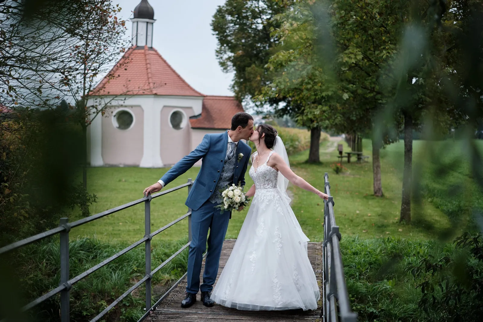 Brautpaar küsst sich auf einer kleinen Brücke vor einer Kapelle – Hochzeit Christina & Manuel