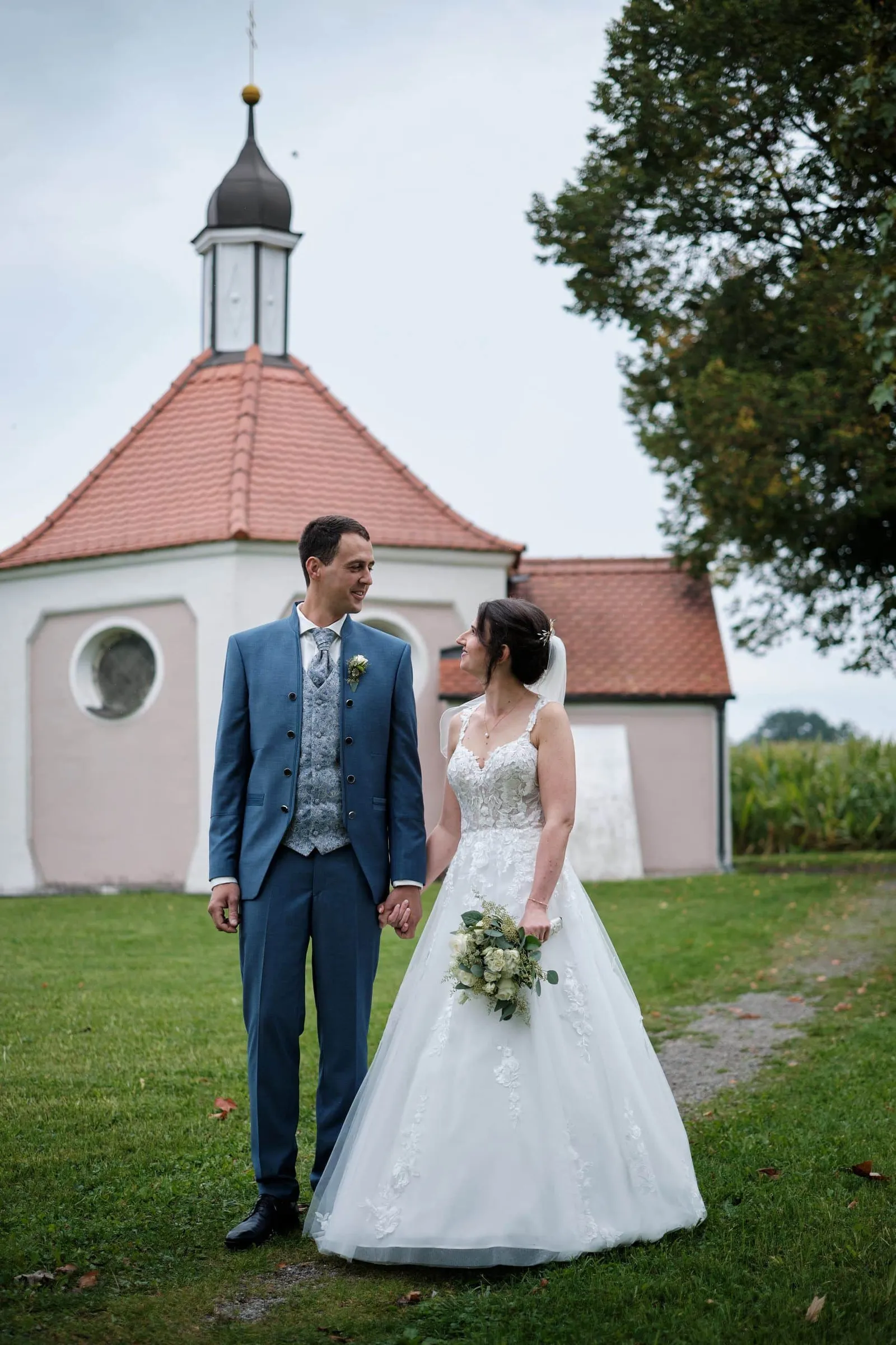 Brautpaar steht Hand in Hand vor einer Kapelle – Hochzeit Christina & Manuel