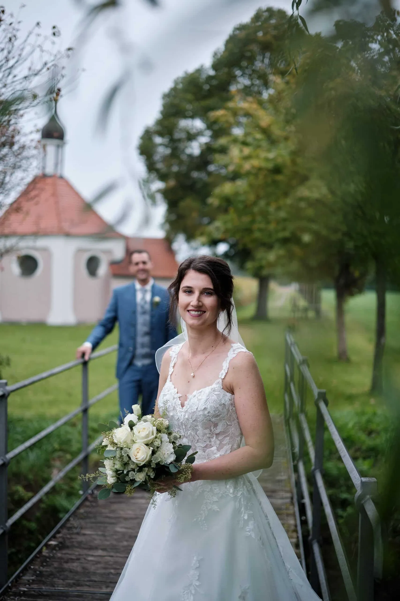 Braut mit Brautstrauß auf einer Brücke, Bräutigam im Hintergrund – Hochzeit Christina & Manuel