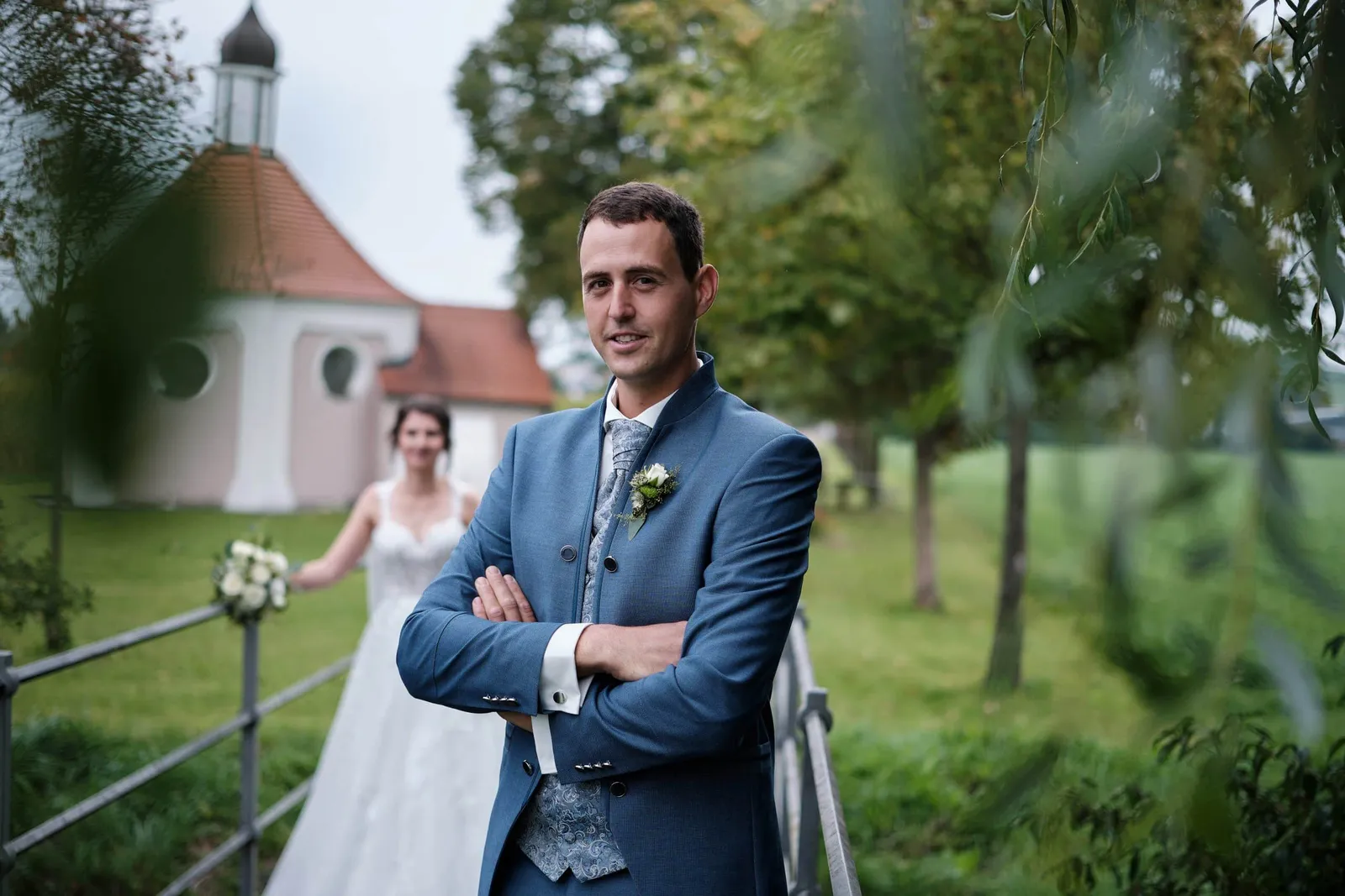 Bräutigam mit verschränkten Armen auf der Brücke, Braut im Hintergrund – Hochzeit Christina & Manuel